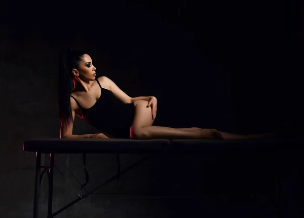 Verleidelijk jong sexy brunette vrouw in zwart bodysuit is liggend op massage tafel, aanraken van haar glad zijdezachte huid — Stockfoto