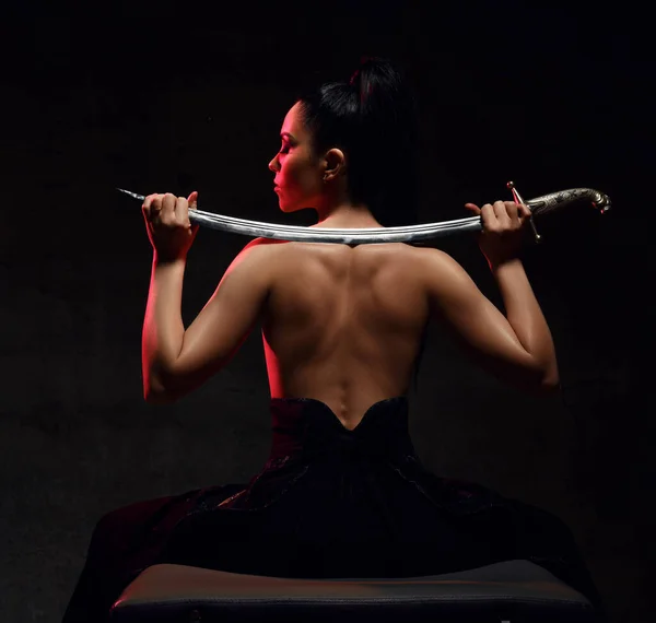 Young brunette woman warrior, martial arts fighter sits topless back to camera holding sword on shoulders, looking aside — Foto Stock