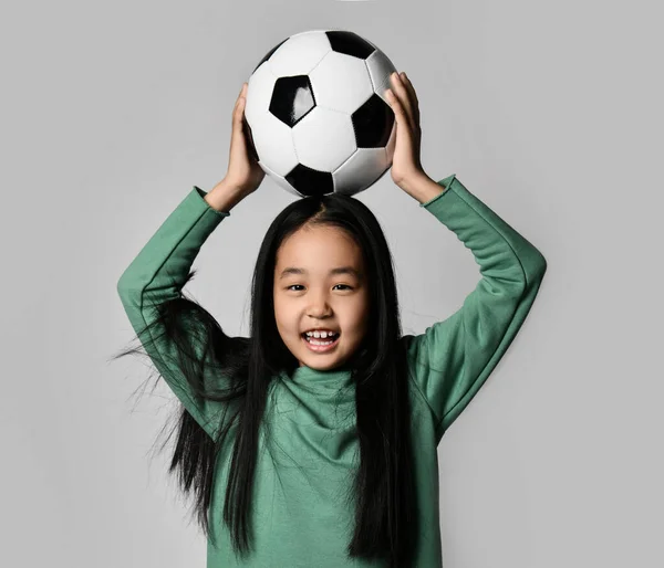 Young beautiful asian girl holding soccer football ball over her head on gray background. Happy Korean soccer player kid — Stock Photo, Image