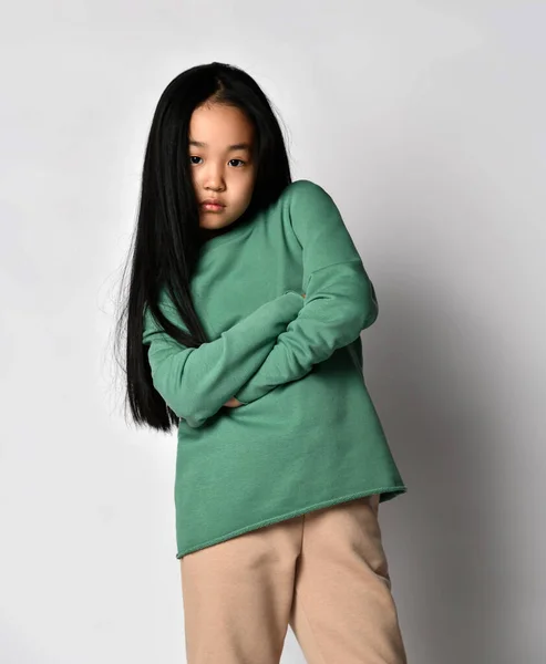 Sensual niña asiática ropa deportiva verde pantalones y sudadera con capucha, suéter se levanta sosteniendo los brazos cruzados en el pecho —  Fotos de Stock
