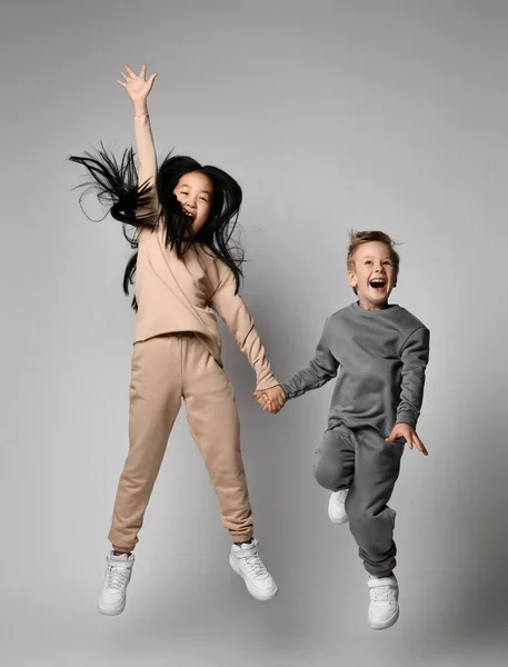 Asian kid girl and caucasian kid boy friends in stylish sportswear have fun jumping and screaming together