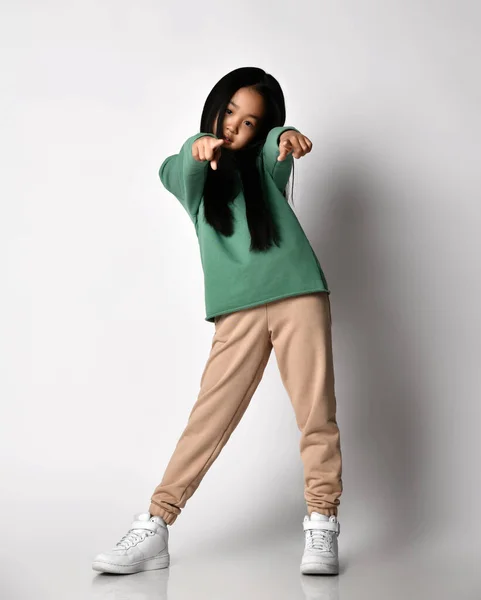 Menina asiática em sapatilhas, calças verdes e capuz, camisola fica apontando com os dedos de ambas as mãos para a câmera — Fotografia de Stock