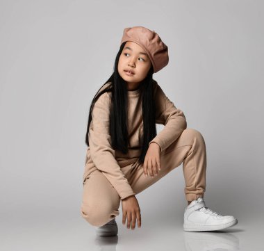 Deri şapkalı, pamuk pantolonlu, kapüşonlu, kazaklı ve spor ayakkabılı Asyalı esmer kız yan yana oturur.