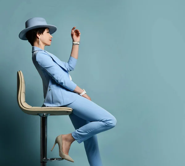 Atraktivní úsměv krátkovlasá brunetka žena v modrém byznysu elegantní ležérní oblek, boty, klobouk sedí na stoličce vypadá rovně — Stock fotografie
