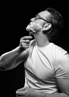 Siyah ve beyaz. Beyaz tişörtlü, gözlüklü, tıraş bıçaklı ve tıraş jeli köpüklü yetişkin bir adamın portresi.