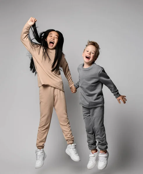 Brunette kid girl and blond kid boy friends in hoodies, pants and sneakers have fun jumping and screaming together