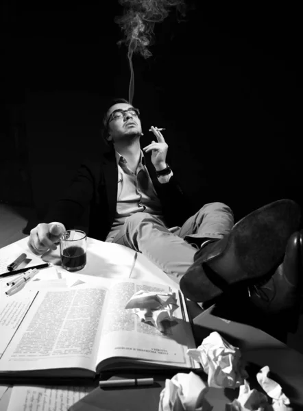 Uomo adulto in abbigliamento casual e occhiali si siede con i piedi sul tavolo con libro e carte, bevande tè, caffè e fumo — Foto Stock