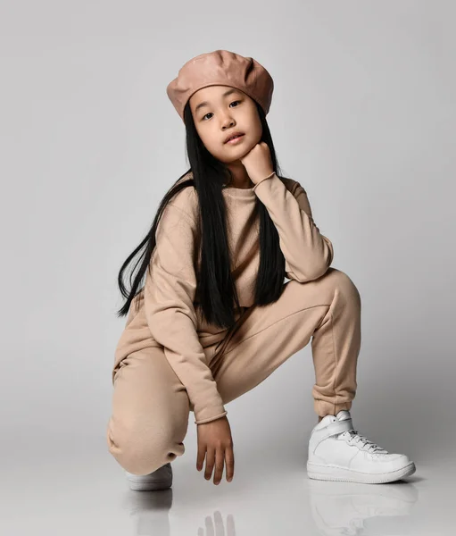 Deri şapkalı, pamuklu pantolonlu, kapüşonlu, kazaklı ve spor ayakkabılı Asyalı esmer kız çömelmiş oturuyor. — Stok fotoğraf