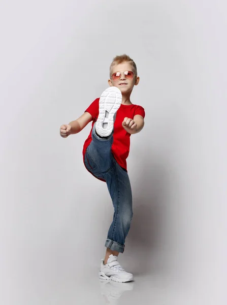 Stylový veselý chlapec dítě v červeném tričku, džíny a sluneční brýle dělat kop s nohou při pohledu do kamery — Stock fotografie