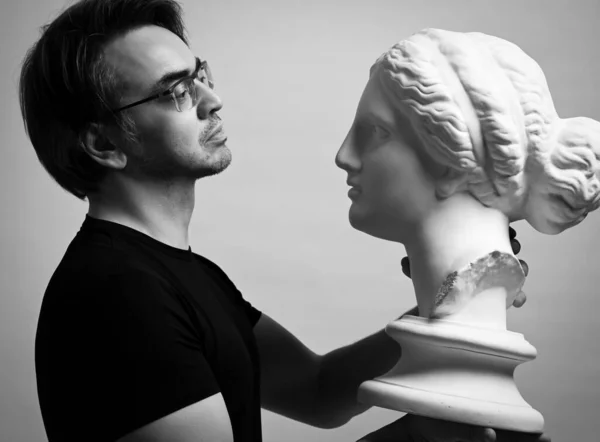 Black and white portrait of adult man in black t-shirt holding in hands, looking at antique sculpture woman head