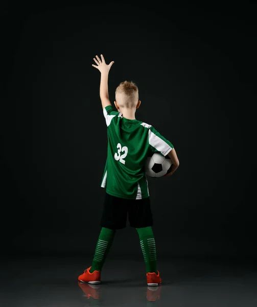 Loira sorrindo menino criança em esportes roupas verdes e brancas de pé segurando bola de futebol e levantou a mão para cima — Fotografia de Stock
