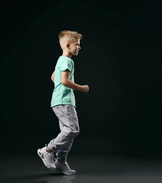 Usmívající se blonďatý chlapec dítě ve sportu zelené tričko, kalhoty a tenisky běží přes tmavé pozadí — Stock fotografie