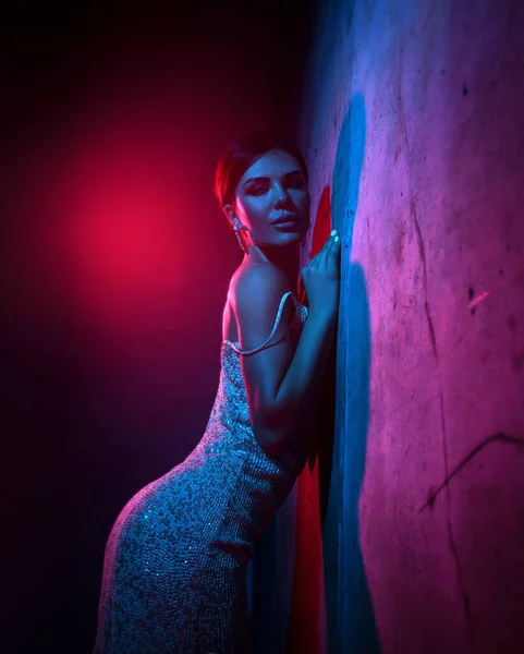 Sexy attractive young adult woman in silver tight cocktail dress is standing at concrete wall in neon light — Stock Photo, Image