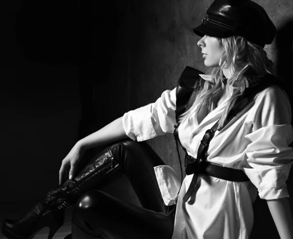 Black and white. Profile of blonde woman in leather pants, white shirt with leather belts and cap sits at concrete wall — Stock Photo, Image