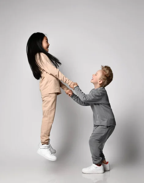 Дети в уютной хлопчатобумажной спортивной одежде играют вместе. Смеющийся блондин мальчик стоит, держа за руки брюнетку девочка прыгает — стоковое фото