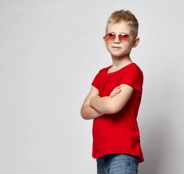 Niño niño con estilo en camiseta roja, jeans y gafas de sol de pie con las manos cruzadas mirando a la cámara —  Fotos de Stock