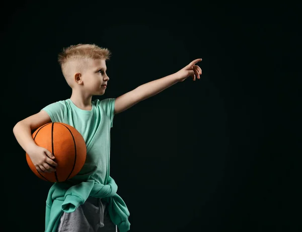 Biondo sorridente ragazzo bambino in sport verde t-shirt e pantaloni in piedi che tiene la pallacanestro in mano rivolta verso l'alto con il dito — Foto Stock