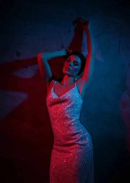 Excited young woman in silver strapless tight cocktail dress is standing at concrete wall in neon light with hands up — Stock Photo, Image