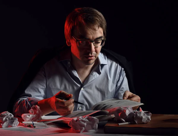 Homem adulto em camisa, jaqueta e óculos se senta atrás da mesa, mesa com lençóis e papel amassado, livro de leitura, escrita — Fotografia de Stock