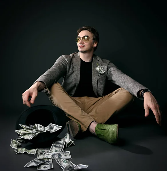 Uomo adulto sicuro di sé in giacca, jeans e scarpe siede gambe incrociate sul pavimento da capovolto cappello pieno di dollari in contanti — Foto Stock