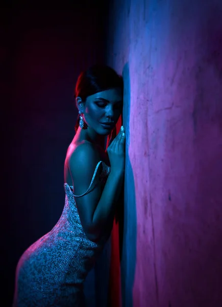 Excited young adult woman in silver tight cocktail dress is standing leaning at concrete wall in neon light — ストック写真