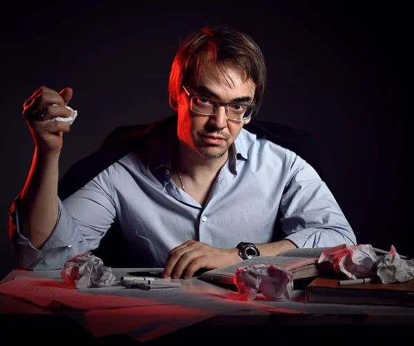 Unshaved pazzo uomo in camicia, giacca e occhiali siede dietro la scrivania, tavolo con libri e lenzuola e carta spiegazzata in pugno — Foto Stock