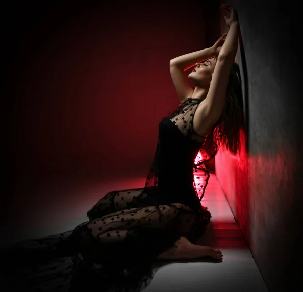 Чувственная молодая блондинка танцовщица в прозрачном кружевном платье и купальнике сидит на коленях на полу у стены с закрытыми глазами — стоковое фото