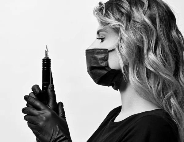 Black and white profile of curly woman in black protective medical mask and gloves holding permanent makeup machine — Stock Photo, Image