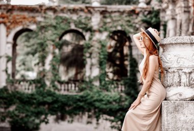 Güneş gözlüklü, şapkalı ve elbiseli şehvetli kadın sanatçı sırtını antik saray bahçesinde taş sütunlara yaslıyor.