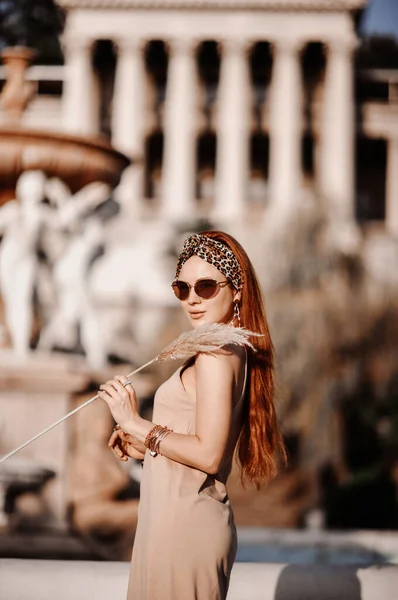 Jonge prachtige roodharige vrouw in zonnebril en zomerjurk staat zijwaarts in de oude tuin van het oude paleis bij fontein — Stockfoto