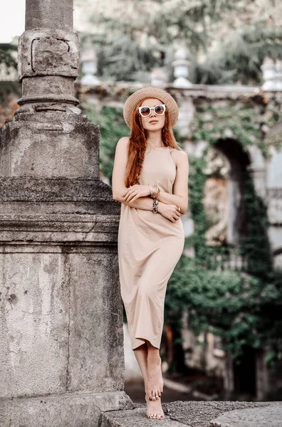 Arrogant woman in sunglasses, hat and dress stands with bare feet on stone parapet at column in ancient palace garden