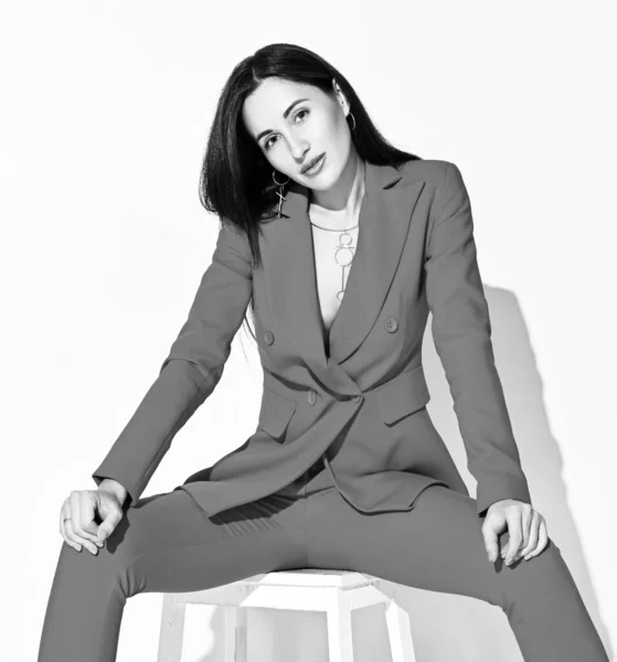 Чорно-білий портрет молодої брюнетки в офіційному костюмі, що сидить на табуреті з розтягнутими ногами — стокове фото