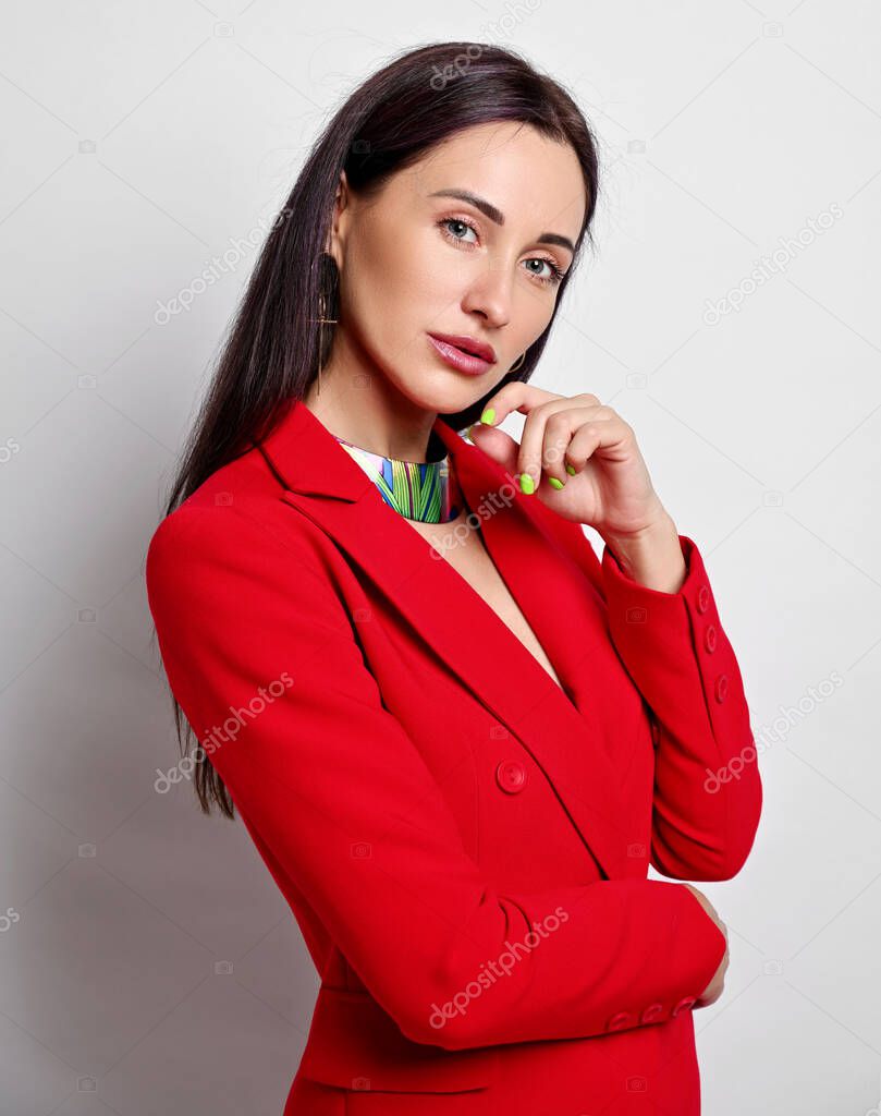 Portrait of businesswoman with permanent makeup in red jacket and modern jewelry standing sideways, looking at camera