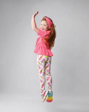 Mutlu, kızıl saçlı, pembe tişörtlü, renkli pantolonlu, sandaletli kız kameraya döndü, zıplıyor ve bize bakıyor.