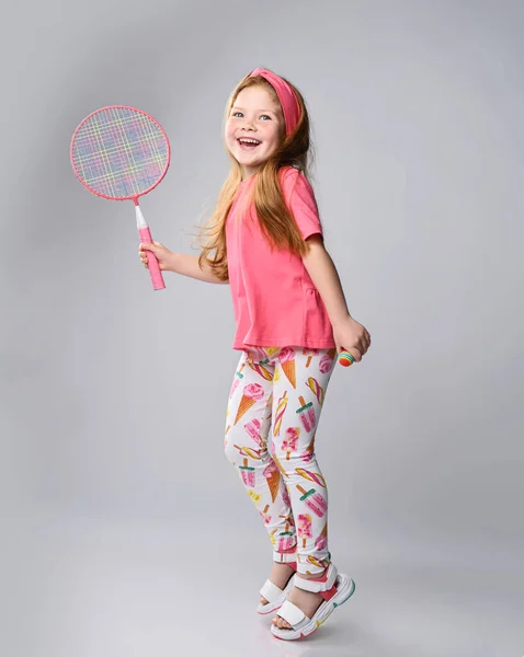 Ativo, rindo menina de cabelos vermelhos em camiseta rosa e saltos de cabeça segurando badminton raquete na mão, joga ao ar livre — Fotografia de Stock