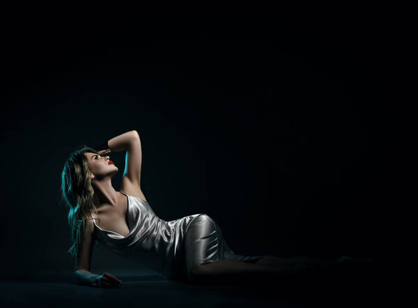 Sexy gorgeous sensual blonde woman in silk silver dress with deep neckline is lying on floor propped up on one elbow looking up in darkness at copy space, holding hand at her head at night