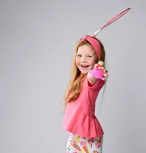 Petite fille aux cheveux roux riante en t-shirt rose et bandeau joue au badminton, tient la navette à la main, raquette balançoire — Photo