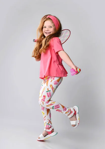 Active red-haired kid girl in pink t-shirt and colorful pants is walking, jumping with badminton racquet on shoulder