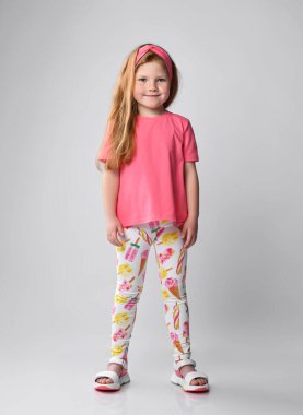 Sevimli, gülümseyen, kızıl saçlı kız yazın ayakta duruyor pembe tişört giyiyor, renkli pantolon giyiyor ve sandalet giyiyor.