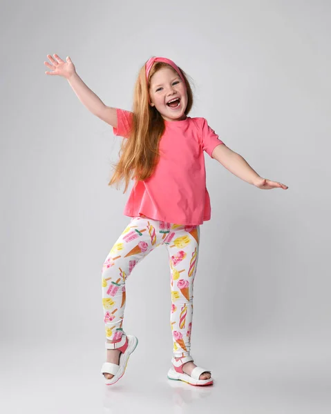 Menina criança ruiva ativa feliz em camiseta rosa, calças coloridas e sandálias fica acenando as mãos, dançando, jogando — Fotografia de Stock