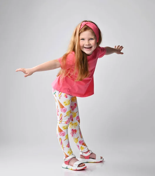 Enfant fille rousse en t-shirt rose, pantalon coloré et sandales danse, joue, se tient la main écartée — Photo