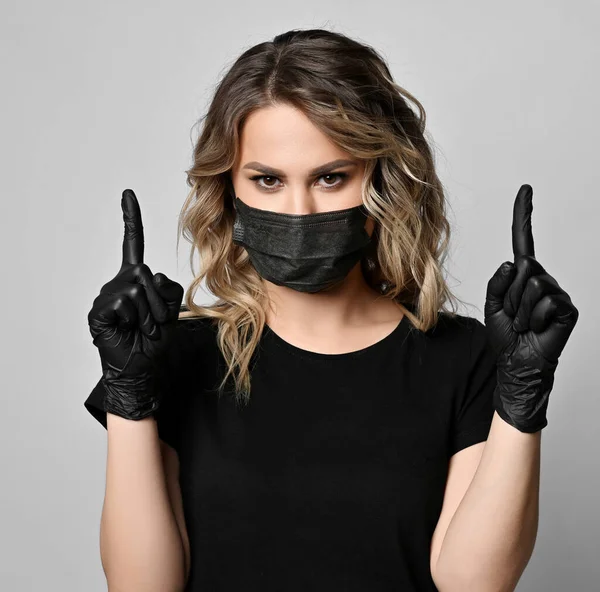 Ritratto di donna riccia bionda in t-shirt nera, maschera e guanti di lattice in piedi tenendo entrambe le mani indice in alto — Foto Stock