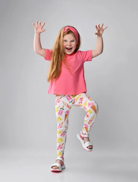Jeune fille rousse espiègle en t-shirt rose, pantalon coloré et sandales joue au dino, monstre tenant la main vers le haut — Photo