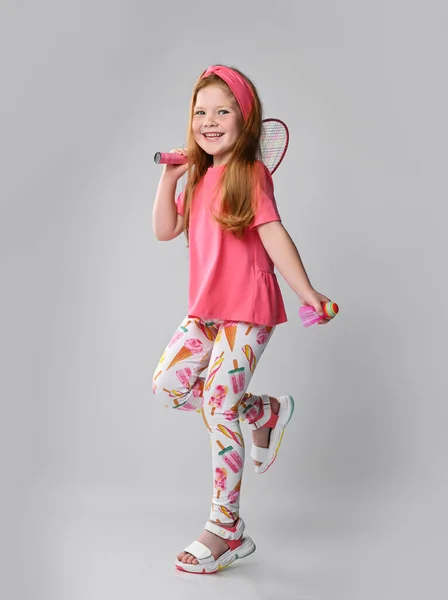 Menina criança ruiva feliz em camiseta rosa, calças coloridas e sandálias está andando vai brincar com badminton — Fotografia de Stock