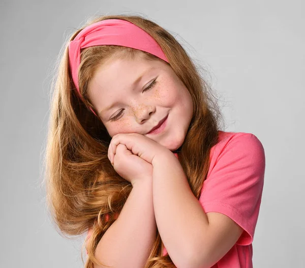 Portrét roztomilé zasněné zrzavé holčičky v růžovém tričku a čelence držící se za ruce se zavřenýma očima — Stock fotografie