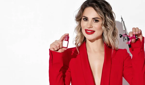 Mujer rubia sonriente en chaqueta roja con escote profundo en cuerpo desnudo sostiene máquina de maquillaje permanente y frasco de pintura — Foto de Stock