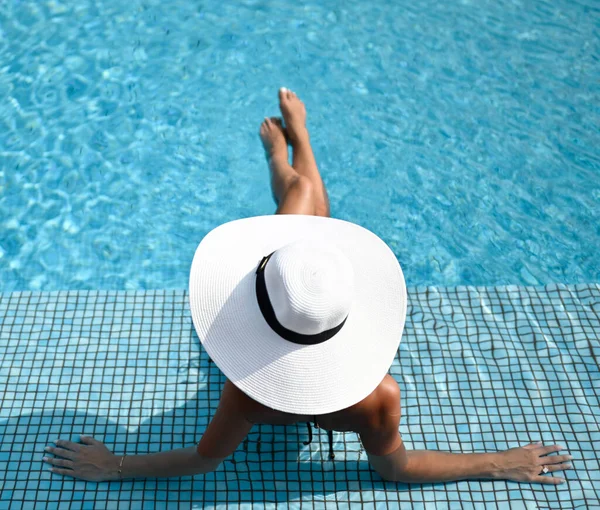 Top view of slim tanned woman in white wide-brimmed hat is sitting in swimming pool clear blue water, relaxing — Stock Photo, Image