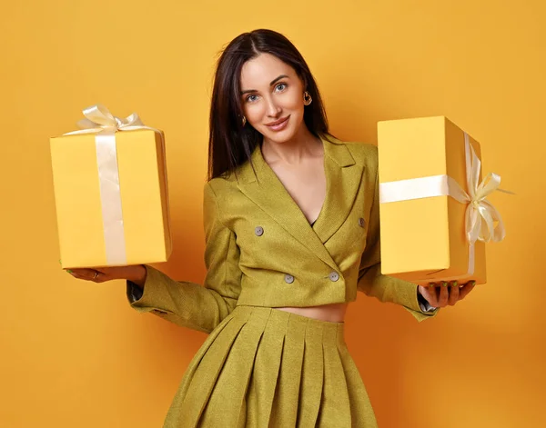 Mooie glimlachende jonge brunette vrouw in geel kostuum rok en jas staande holding aanwezig dozen op zoek naar camera — Stockfoto