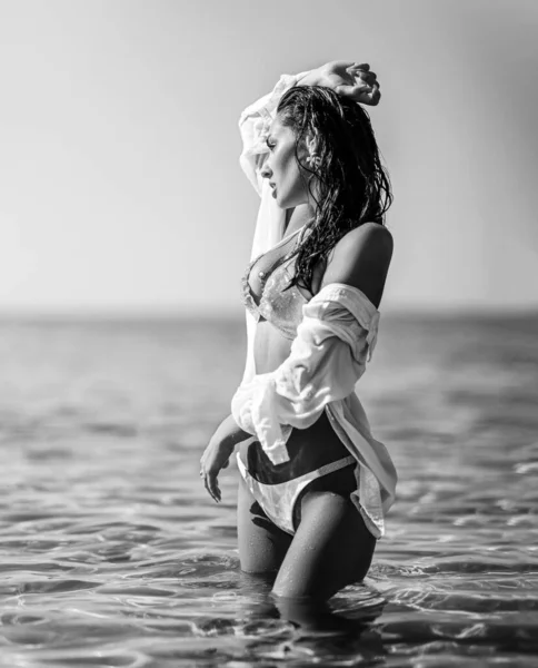 Jonge vrouw in witte bikini en shirt staat in taille-diepe zee, kijkend in de verte. Zwart op wit. Zomer vibes — Stockfoto