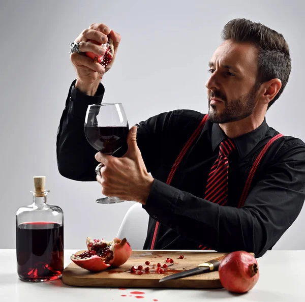 Uomo adulto in camicia nera con cravatta a righe e bretelle si siede a tavola spremendo succo di melograno fresco in un bicchiere — Foto Stock